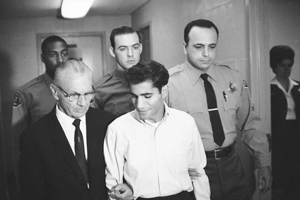 June 1968 photo of Sirhan Sirhan being escorted by law officers and a man in a dark suit