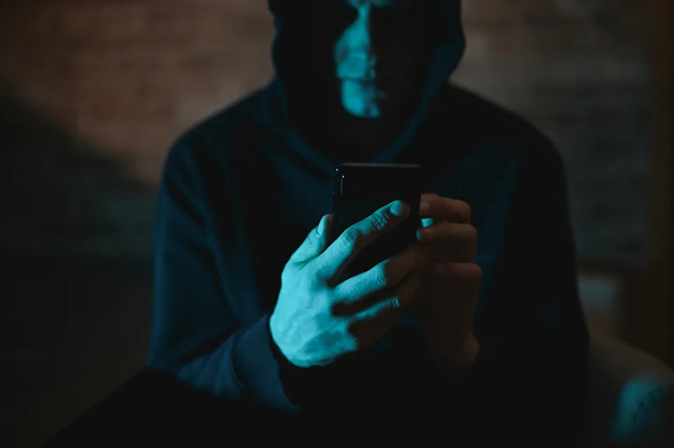 Person in hoodie using a smartphone in a dimly lit setting, suggesting secrecy or focus