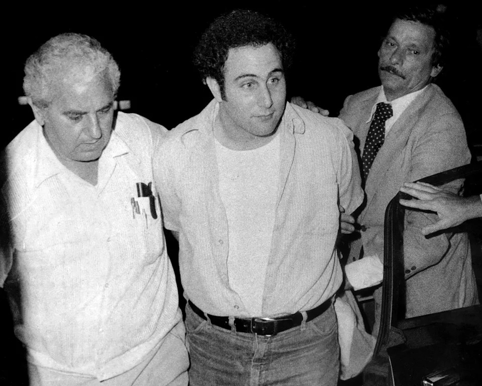 three men walk together, the man in the middle is escorted with his hands behind his back