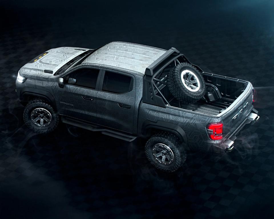Matte gray pickup truck with a geometric pattern on the body, featuring off-road tires and a spare tire mounted vertically in the truck bed. The truck has a double cab, black side steps, and a reinforced roll bar behind the cab. The rear lights are illuminated, and the truck is positioned on a dark, checkered surface with a smoky atmosphere.