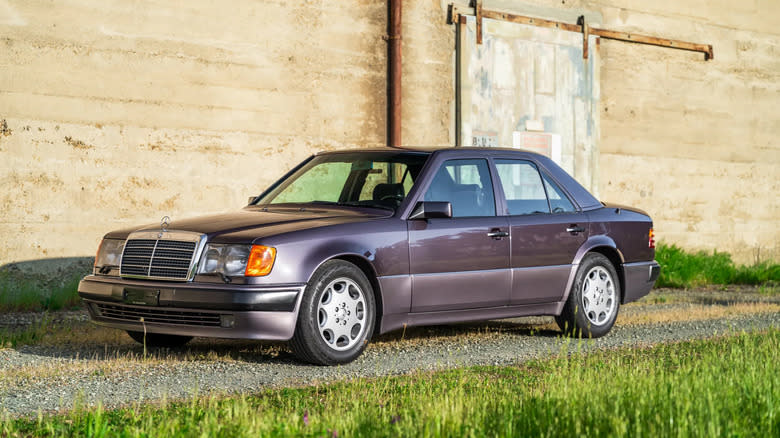 A purple 1992 Mercedes-Benz 500e parked on gravel in front of a wall