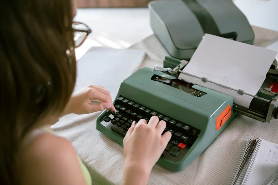A person typing on a vintage typewriter at a table, with notebooks nearby