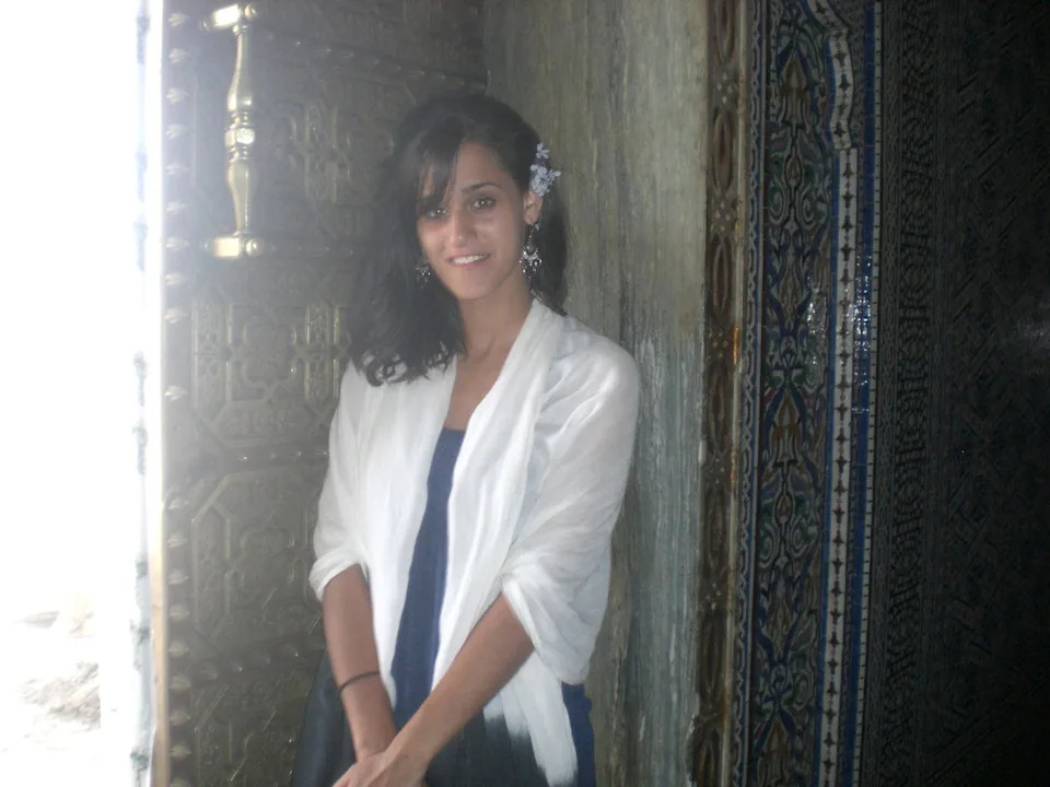 Person standing by an ornate doorway, wearing a shawl over a dress, with flower hair accessory