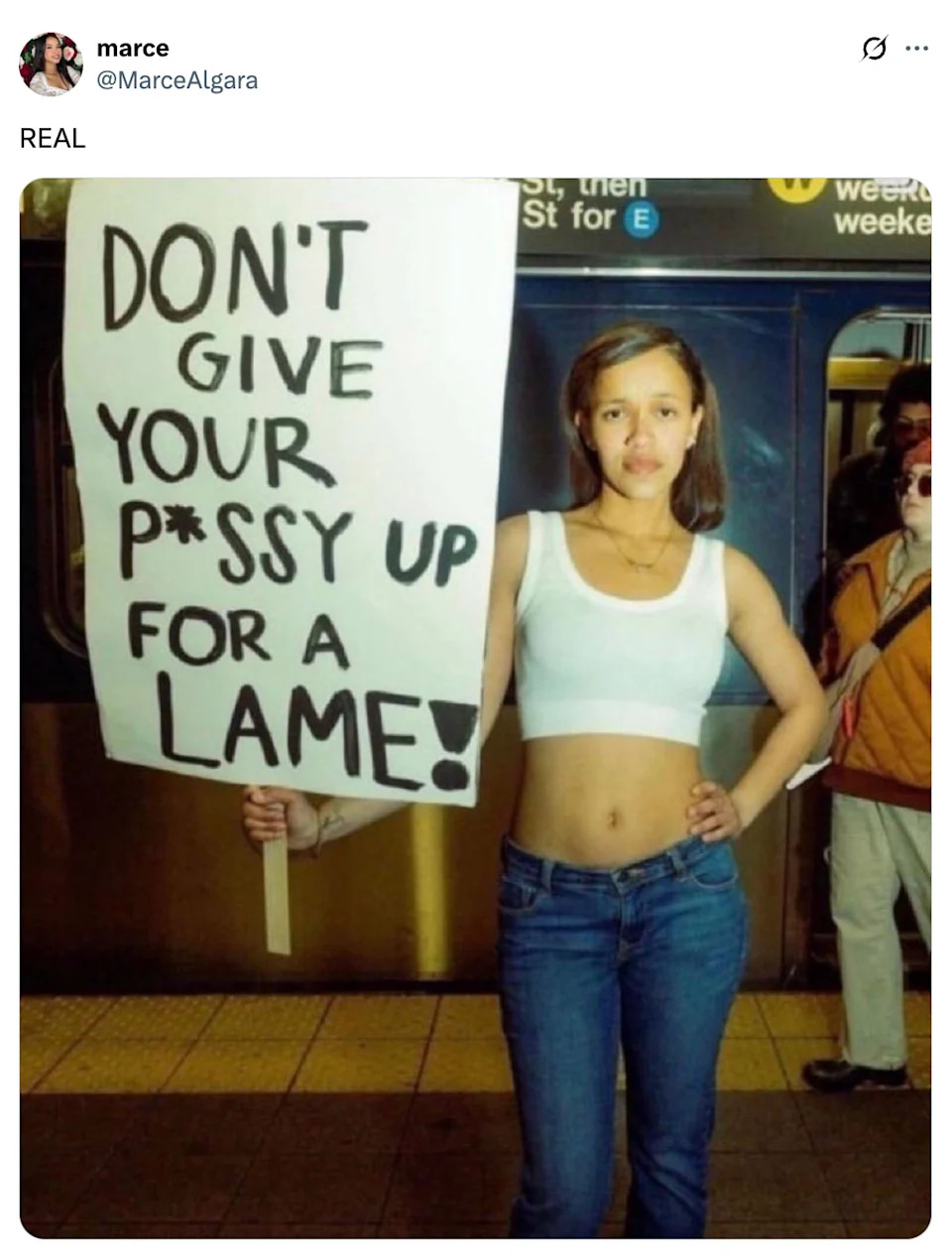 A person in a subway station holds a sign saying, "DON'T GIVE YOUR P*SSY UP FOR A LAME!"