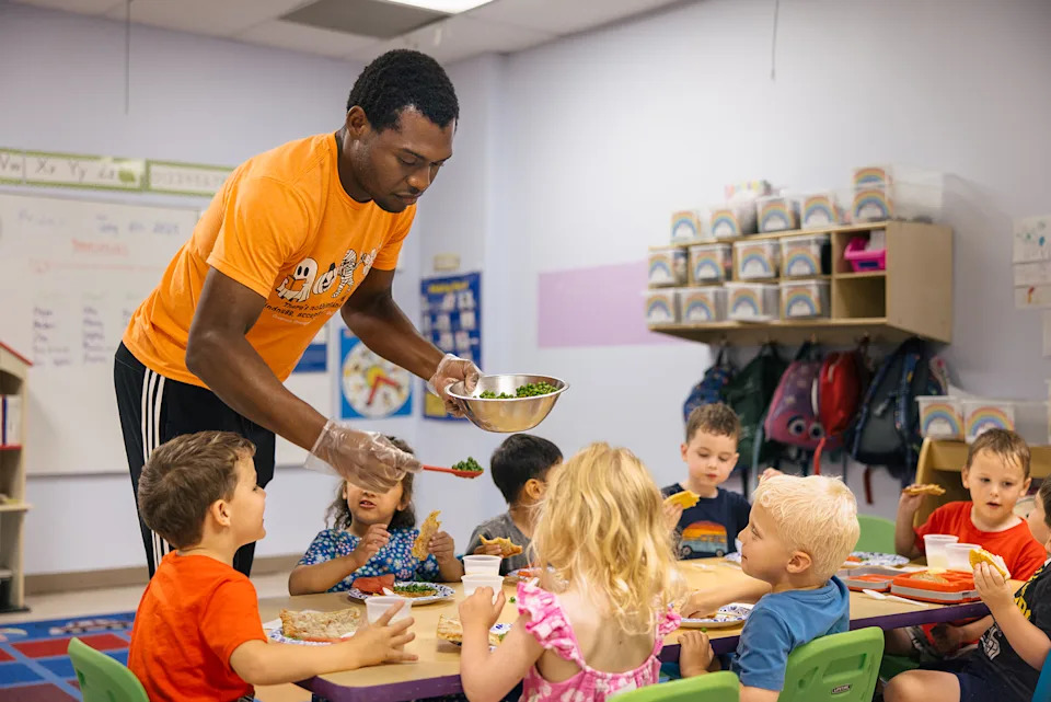 A young man serves peas to a table of young children in a childcare classroom