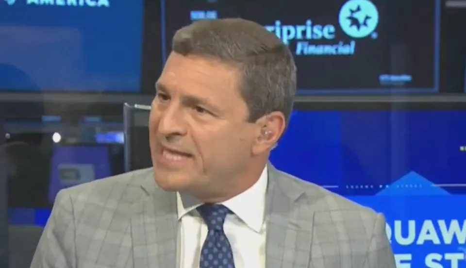 A man in a checked suit speaks during a financial news broadcast, with company logos in the background