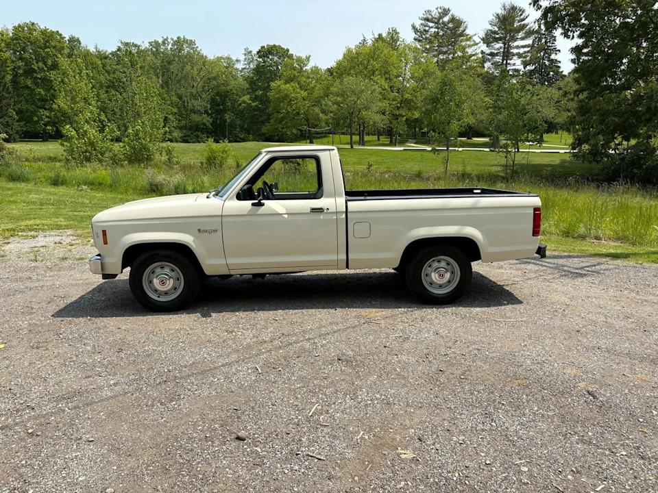 side view of beige pickup truck parked on gravel with green trees and grass in background.