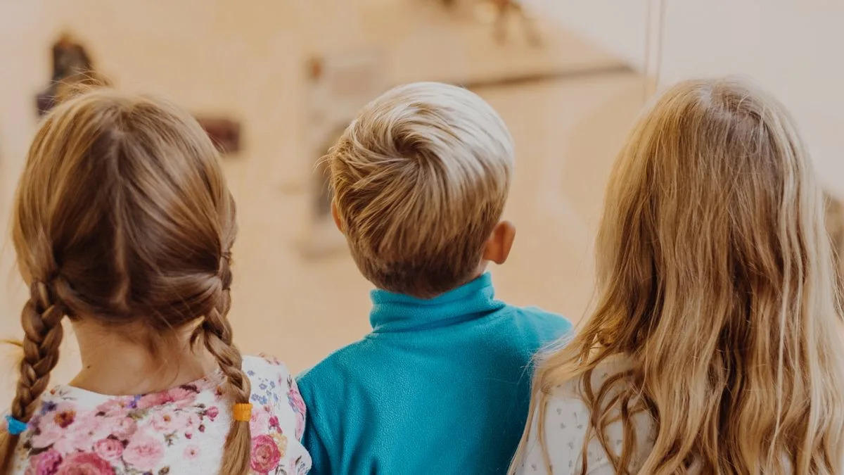 Three young children are seen from behind, standing close together and looking out through a glass window. The child in the center has short blond hair and wears a bright blue fleece, while the two children on either side have long, light brown hair. One wears a floral-patterned shirt and has her hair in braids, and the other has loose hair and wears a light-colored top. 