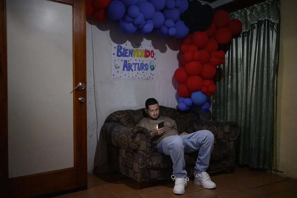 A man with dark hair, seated under a hand-drawn sign and a cluster of red and purple balloons, looks at his phone