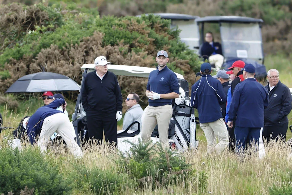 A number of people, mostly men, on a golf course with a number of golf buggies. President Donald Trump and his son Eric are in the centre of the shot