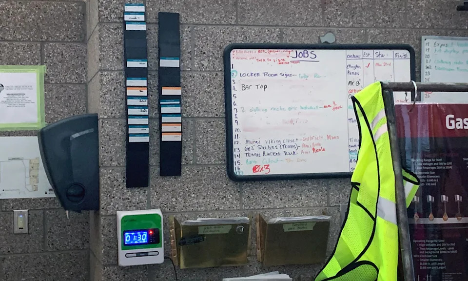 Electronic time clock that welding teacher Shawn Coffey installed in his classroom, with students’ time cards on racks above. At right is a whiteboard where students track the jobs they have solicited around the school, from bid to completion. (Beth Hawkins)