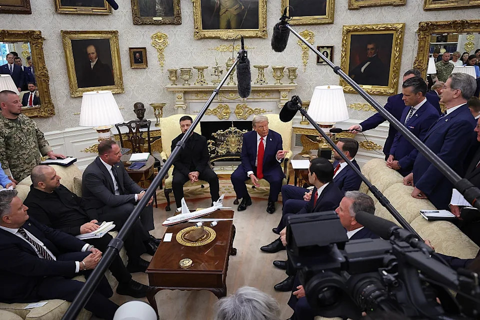 People seated in a meeting surrounded by cameras and microphones, with framed portraits and model planes visible in the background