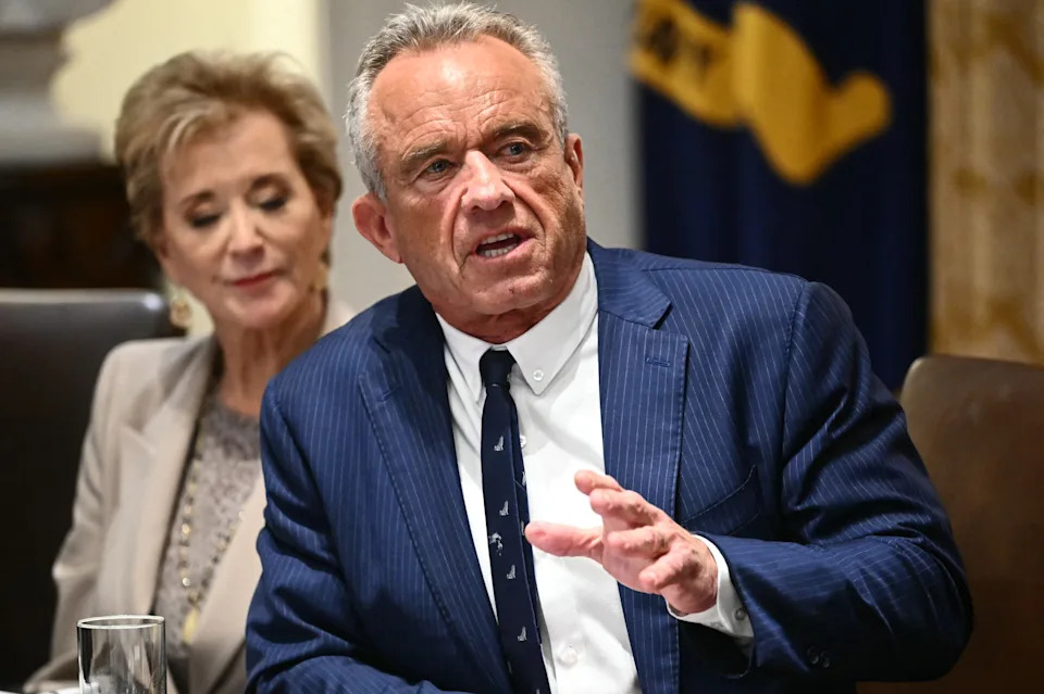 A man in a pinstripe suit gestures while speaking at a formal gathering. A woman sits beside him, listening attentively