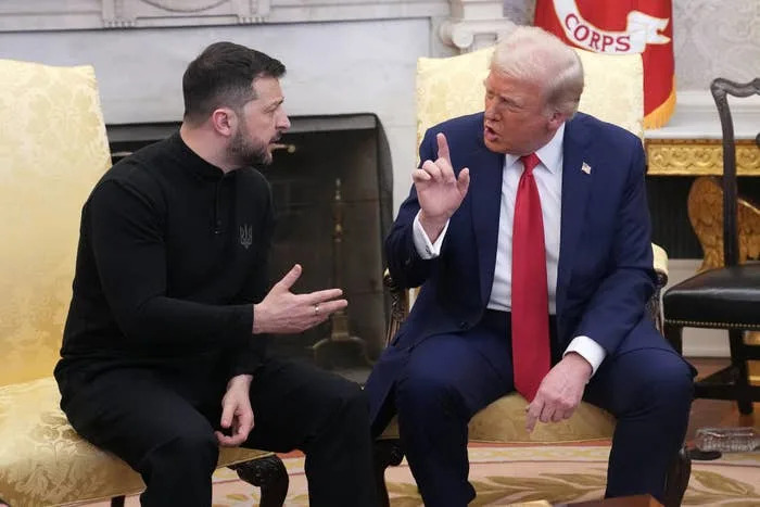 Two men are seated, engaged in a serious conversation. One wears a dark casual outfit, the other a formal suit