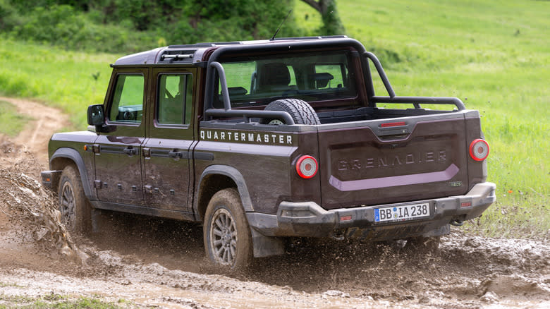 An INEOS Grenadier Quartermster pickup truck in rear view driving though muddy terrain.