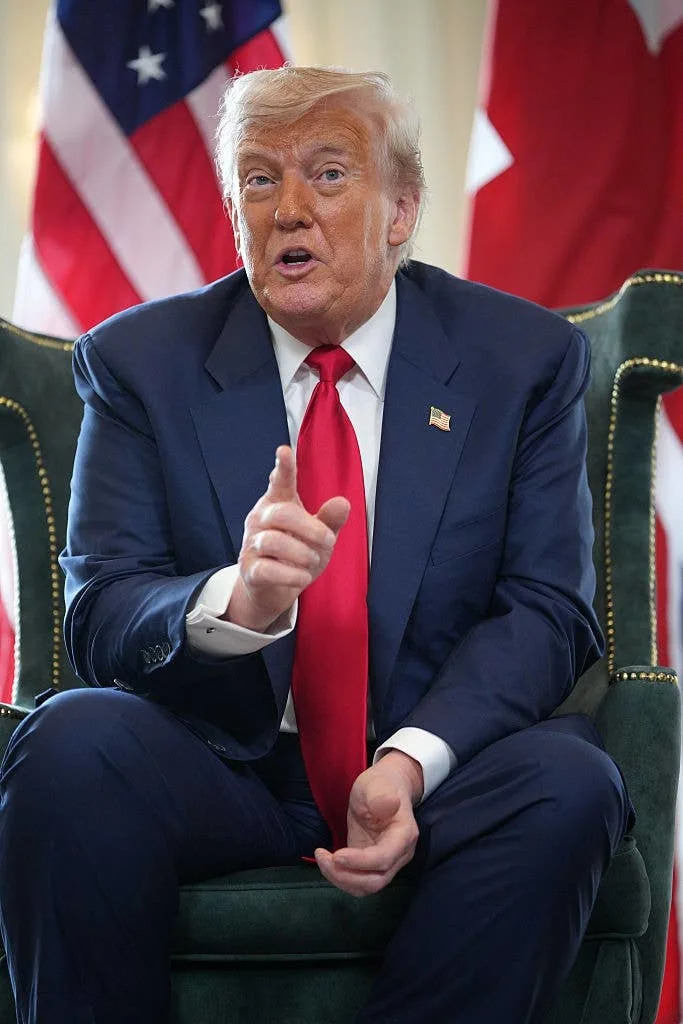 Person in a suit with a red tie sits on a chair, gesturing during a speech. U.S. flag in the background