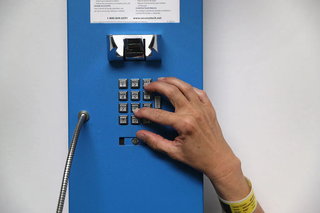 A person's hand is shown dialing an industrial blue push-button phone attached to a wall.