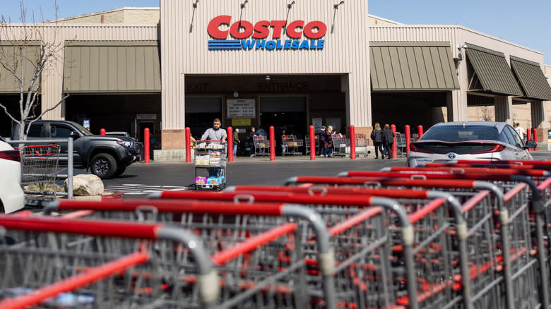 A line of Costco shopping carts being pushed through the parking lot, as shoppers enter and exit the store.