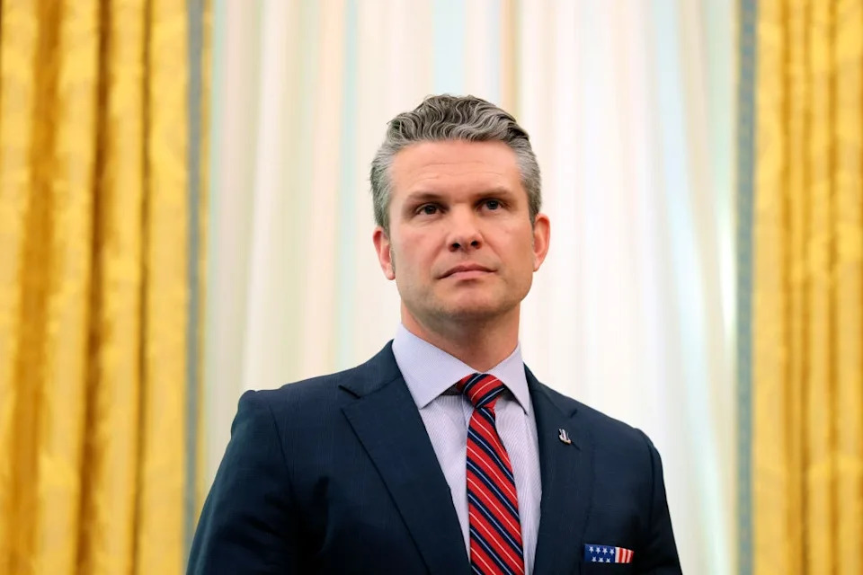 A person in a suit with a striped tie stands in a formal setting. Drapes in the background