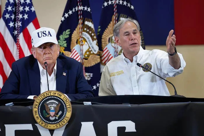 Two men seated at a table with microphones; one in a white shirt gestures while speaking, and the other wears a USA cap and suit
