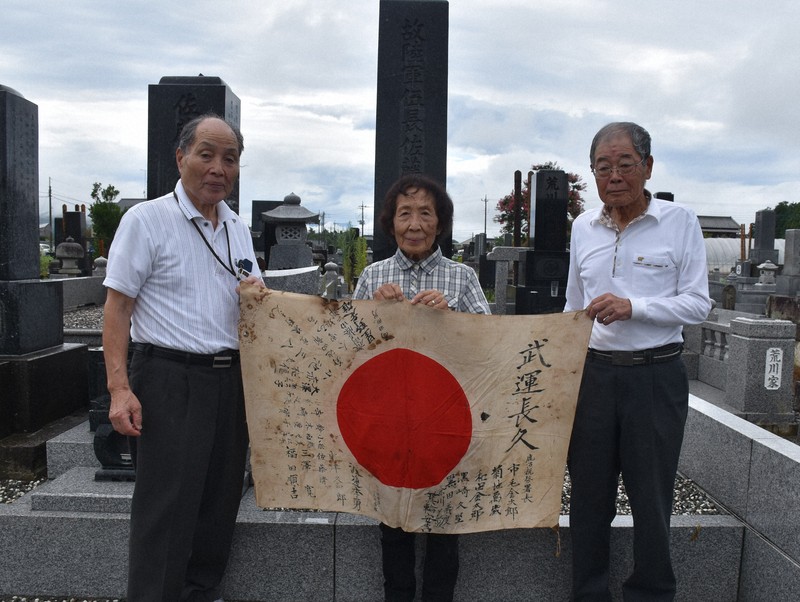 佐藤清さんの墓前で日章旗を広げる稔さん、美惠子さん夫妻と小倉さん(右から)=栃木市西方町で2025年8月7日午前11時1分、太田穣撮影