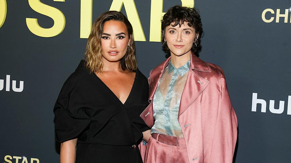 Alyson Stoner con un traje de satén rosa y una blusa de encaje azul claro posando junto a Demi Lovato con un vestido negro escotado.