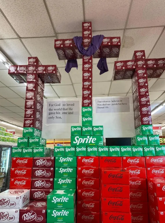 Soda boxes form three crosses in a store, with religious text displays about God's love and belief in His Son nearby