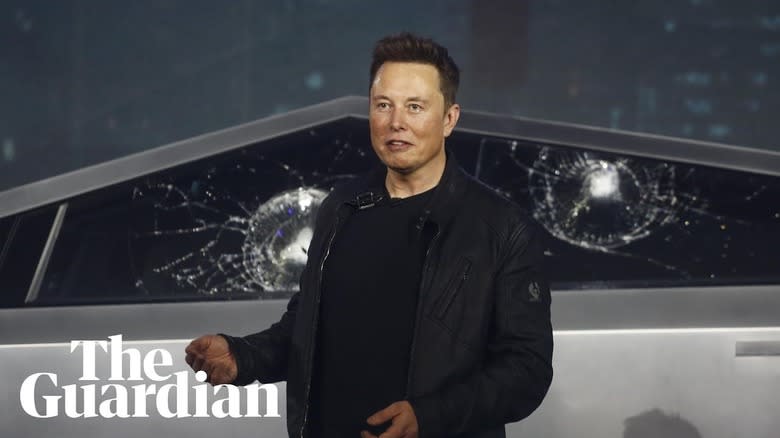 Elon Musk standing in front of a Cybertruck with two shattered windows after a failed demonstration of its shatterproof glass