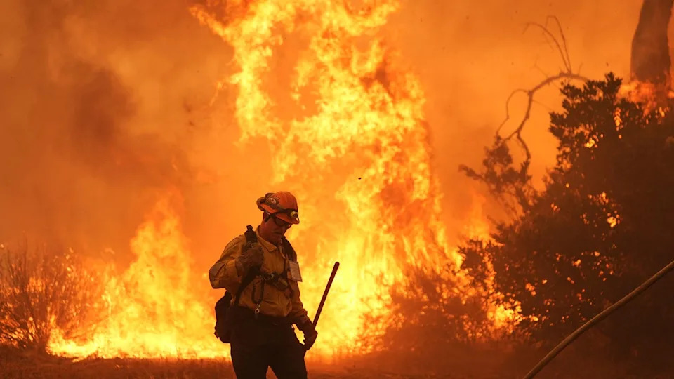 A firefighter looks down at the ground while a large fire burns behind him