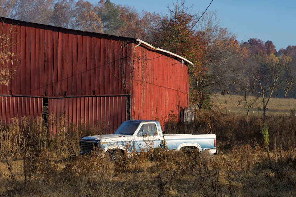 Old pickup truck parked beside a worn, wooden barn in a rural, overgrown field