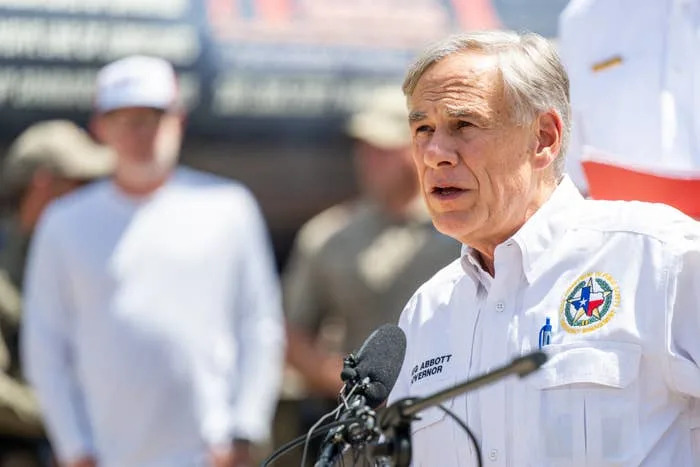 A man in a uniform shirt speaks at a podium with a microphone, delivering a statement at an outdoor event; people and a banner are in the background