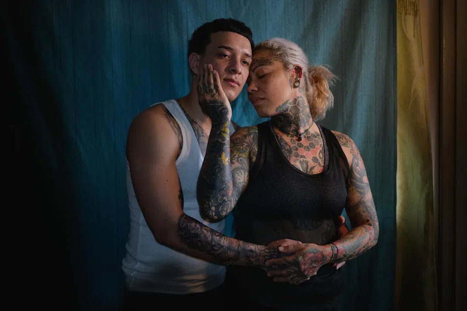 A blond woman covered in tattoos holds the face of a young man, with her other hand over his