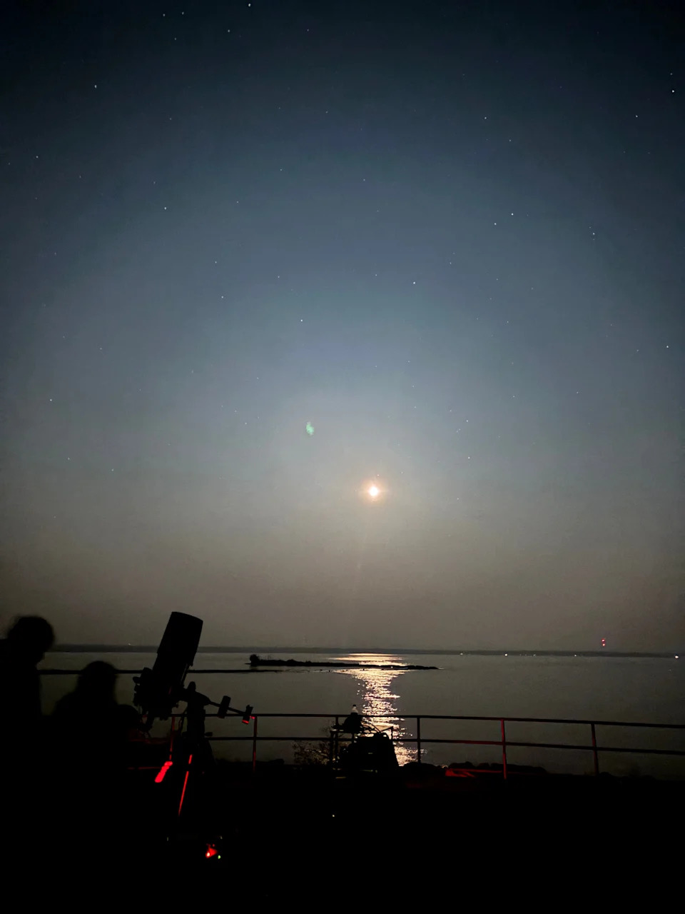 The moon shines over the water at the Headlands International Dark Sky Park while people line up to view a star cluster through a telescope.