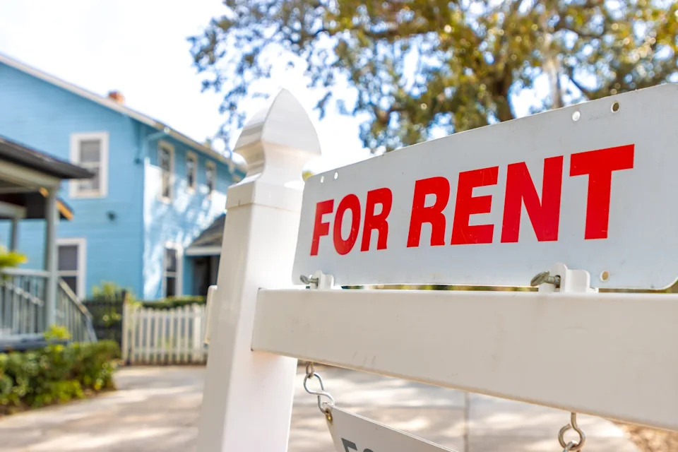 "For Rent" sign in front of a home with a porch and picket fence, suggesting the house is available for lease