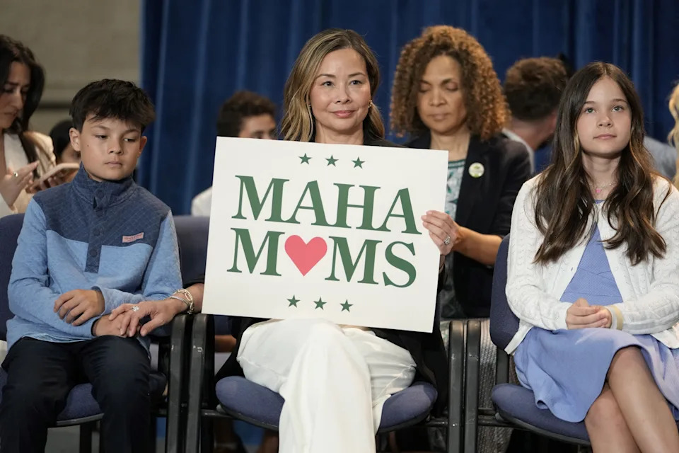 Mothers and children sit in rows of chairs, with one woman smiling and holding a sign reading “MAHA Moms.”