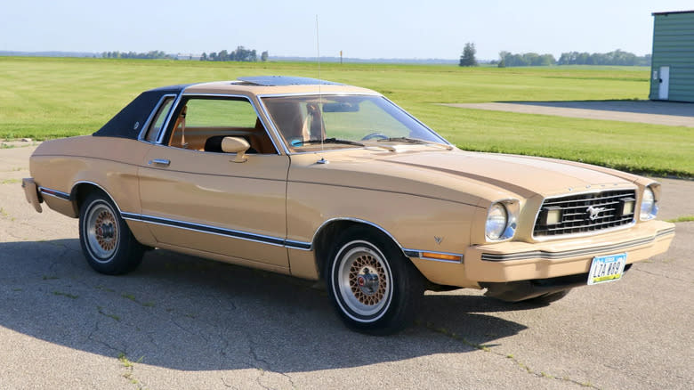 A cream colored Mustang II parked in front of a flat grass field