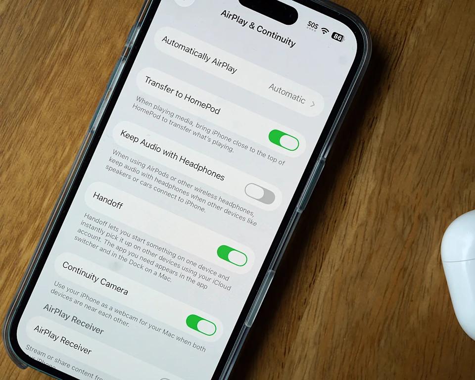 iPhone screen showing the "AirPlay & Continuity" settings menu. Options visible include "Automatically AirPlay" set to Automatic, "Transfer to HomePod" enabled, "Keep Audio with Headphones" disabled, "Handoff" enabled, and "Continuity Camera" enabled. The phone is in a clear case and placed on a wooden surface next to a white AirPods case.