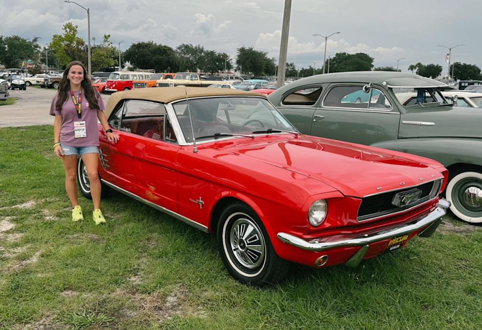 Woman standing proudly next to her 1966 Ford Mustang Convertible at Mecum auction.