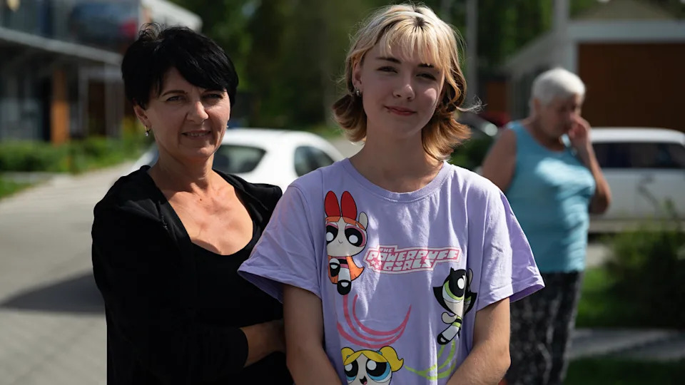 Valeria (right) stands in a lilac Powerpuff Girls t-shirt with her mother in their refugee village