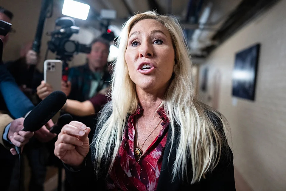 Person with long hair speaking to reporters, wearing a floral blouse and blazer, with cameras and microphones around
