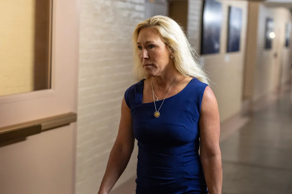 Person walking in a hallway, wearing a sleeveless, fitted dress and a pendant necklace
