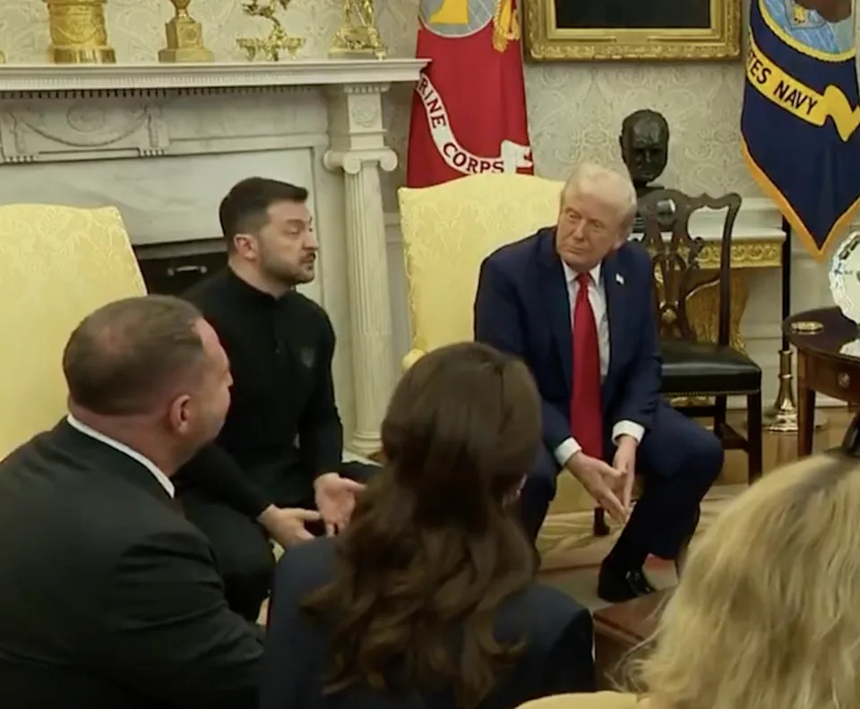 Two men sit talking in a formal room, with others around them. One wears a suit; the other wears a casual outfit. Military flags and decor are visible