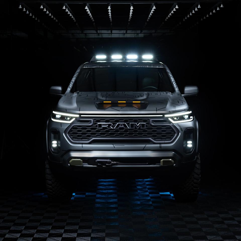 Front view of a silver RAM pickup truck with illuminated LED headlights, roof lights, and fog lights, set against a dark background. The truck features a bold grille with the RAM logo and a hood scoop with amber marker lights. The floor beneath the truck has a checkered pattern with blue lighting accents.