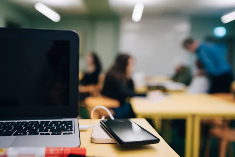 Open laptop and smartphone on a table in a classroom setting, with blurred figures in the background suggesting a casual work environment