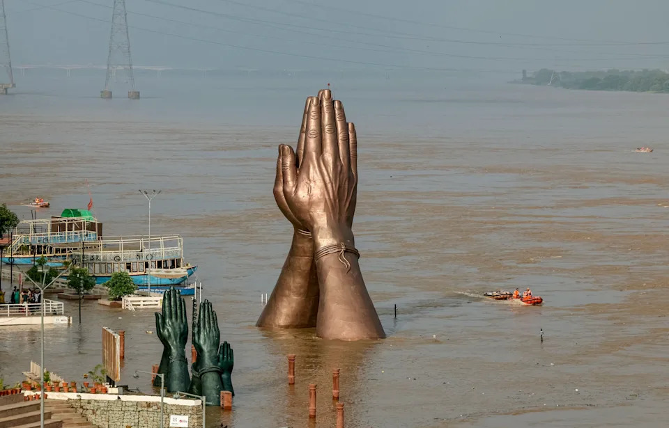 A large statue of hands placed together, seen along a flooded riverbank.