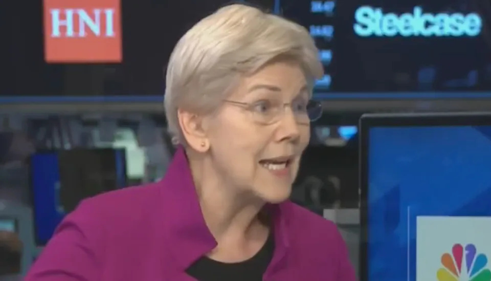 Person speaking animatedly during a televised interview, wearing a purple jacket, with monitors in the background displaying company logos