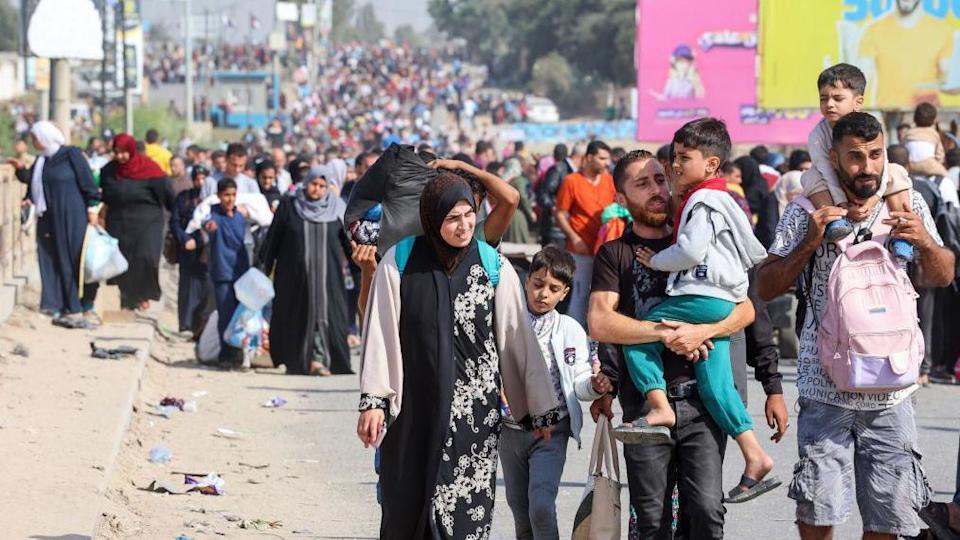 Una multitud de personas camina por una carretera en Gaza el 10 de noviembre de 2023. En primer plano, dos hombres llevan a niños pequeños, uno con un niño a hombros y una mochila rosa atada al pecho. Junto a ellos, una mujer camina junto a un niño mayor. Detrás de ellos, la multitud se extiende por la carretera hasta donde alcanza la vista.