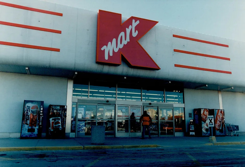 Storefront of a Kmart with large "Kmart" sign above entrance. People are entering beneath the canopy. Vending machines are visible on the left