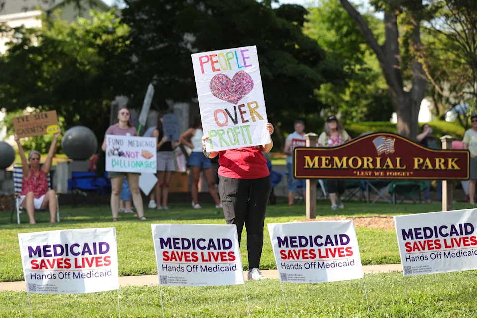 Protester holding "People Over Profit" sign at a Medicaid rally, surrounded by supportive signs and people at a park