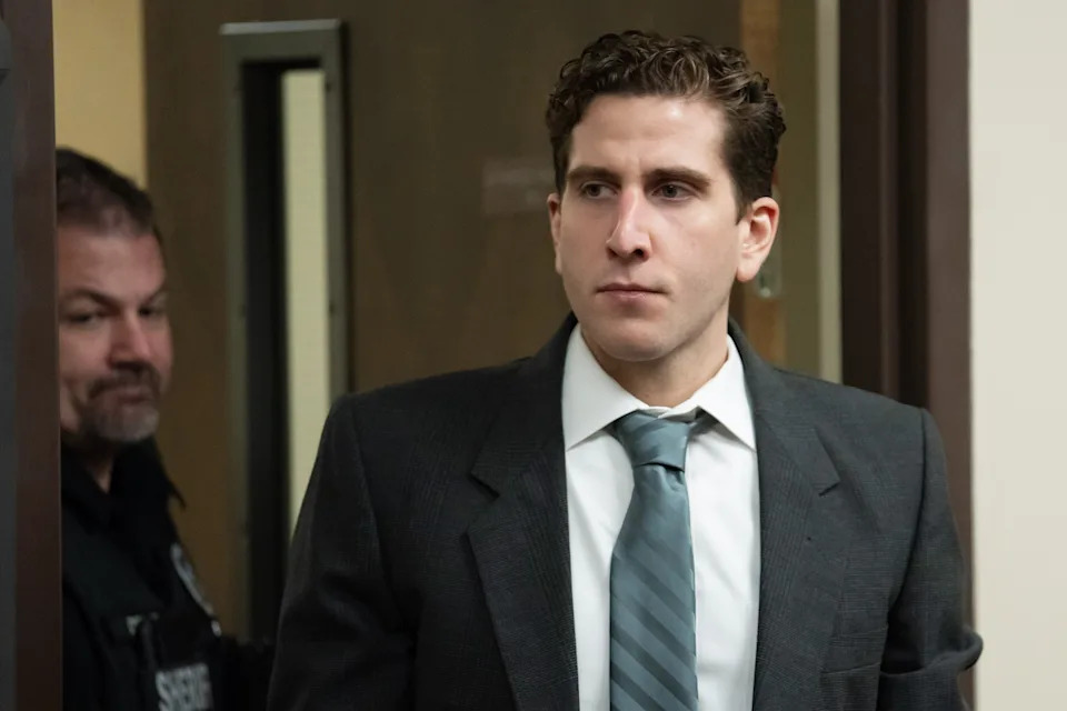 A man in a suit and tie walks through a doorway, followed by a uniformed officer
